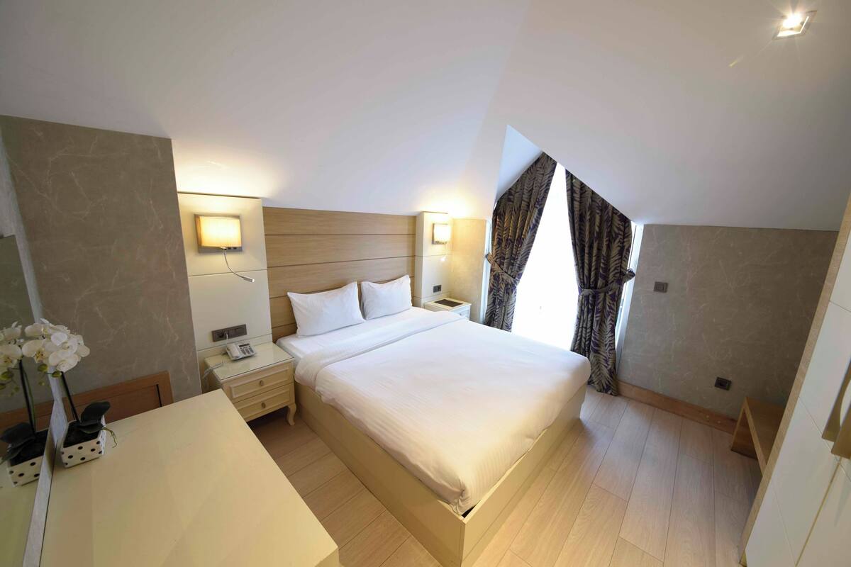 A hotel room with a bed, bedside tables, a desk, and a window with curtains.