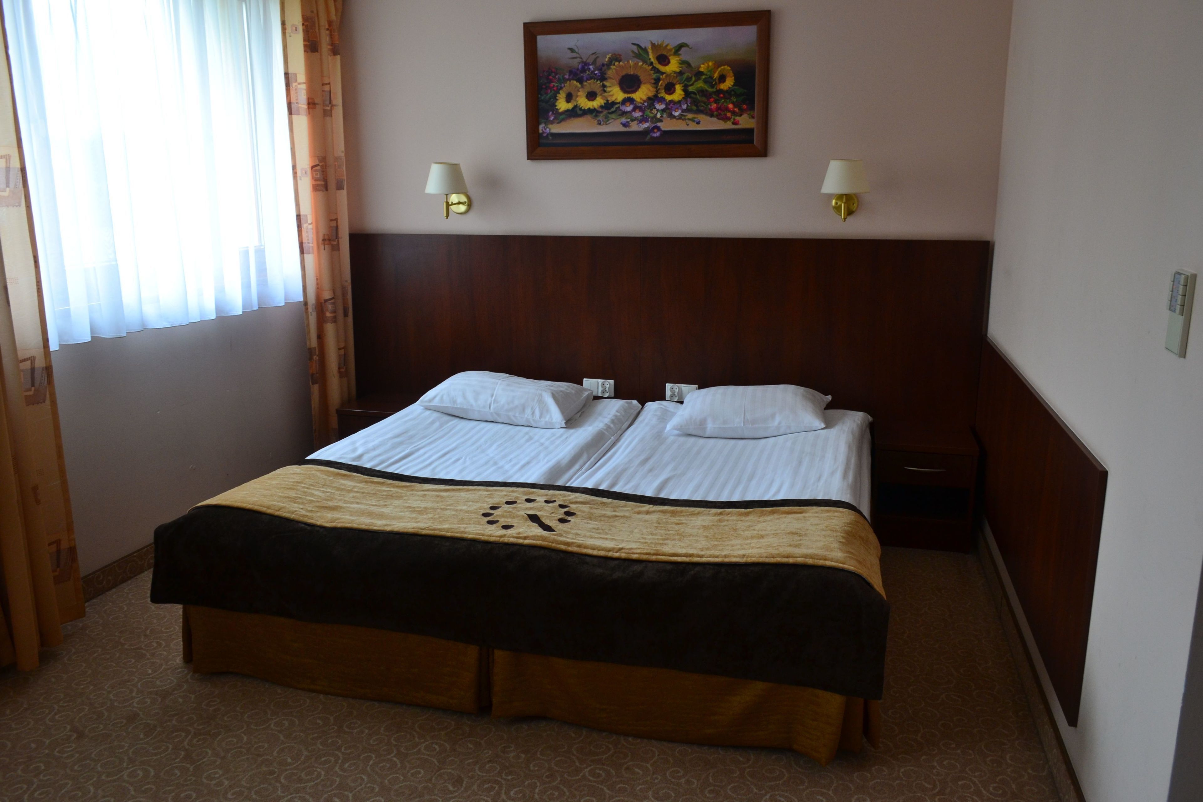 Quarto de hotel com cama de casal, duas mesas de cabeceira, um relógio sobre a colcha, um quadro na parede e duas luminárias fixadas na parede.