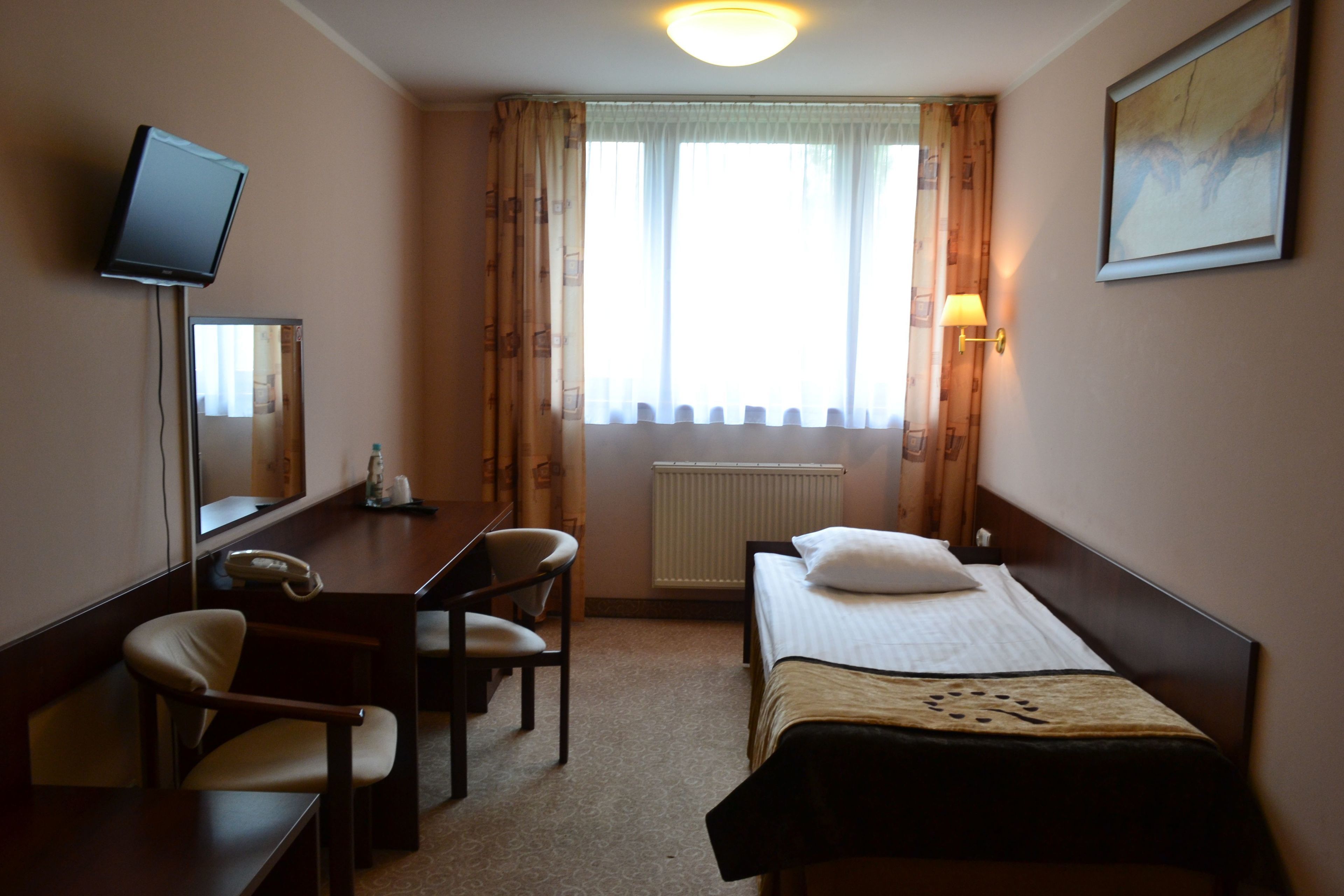 A hotel room with a bed, a desk with a chair, a television mounted on the wall, and a window with curtains.