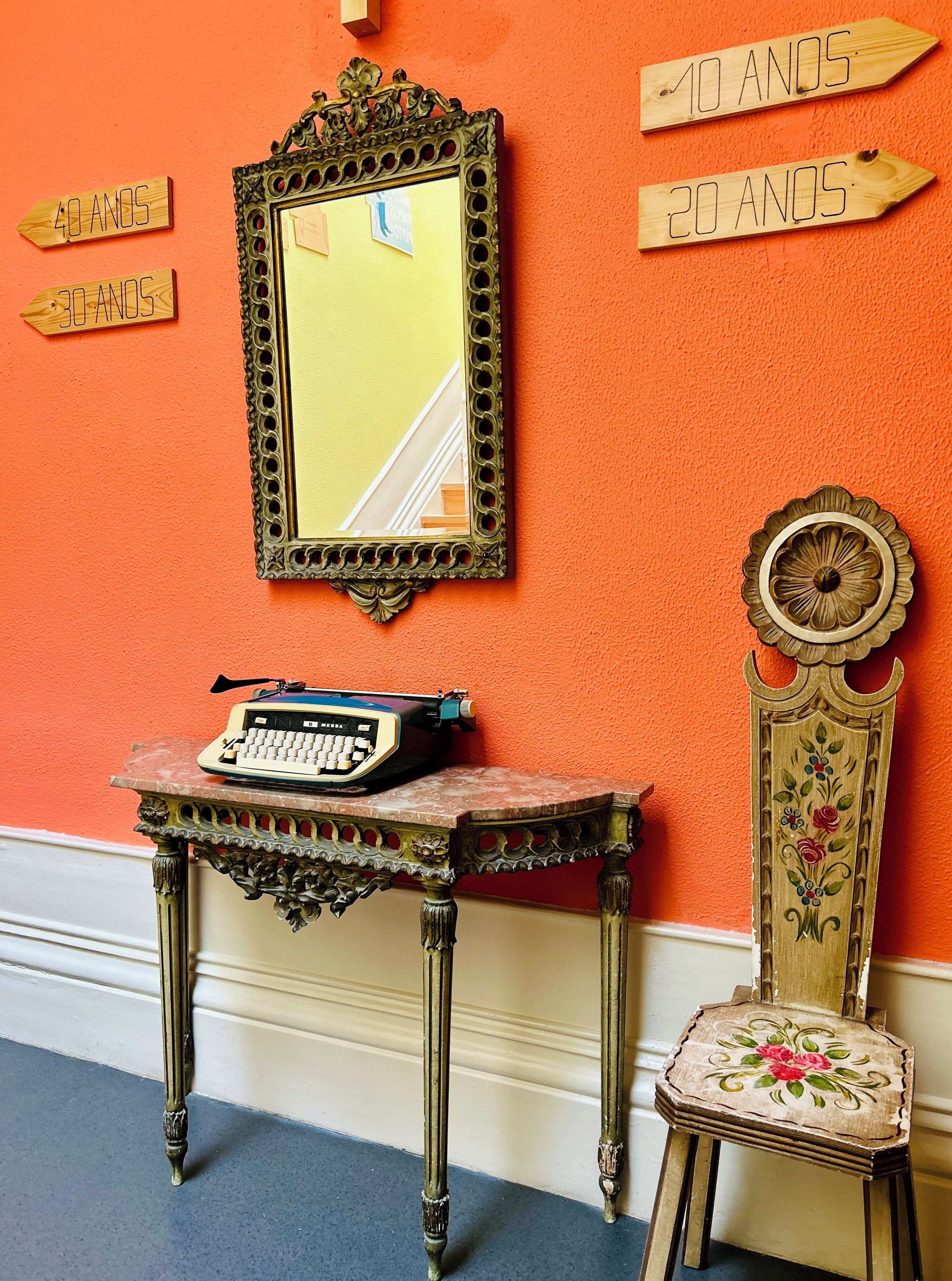 Una mesa de estilo vintage con una máquina de escribir, un espejo decorativo y una silla con tapicería floral frente a una pared naranja.