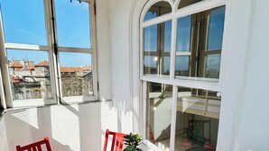 Un balcón con una mesa y sillas rojas, una planta en maceta y vistas de edificios a través de amplias ventanas.