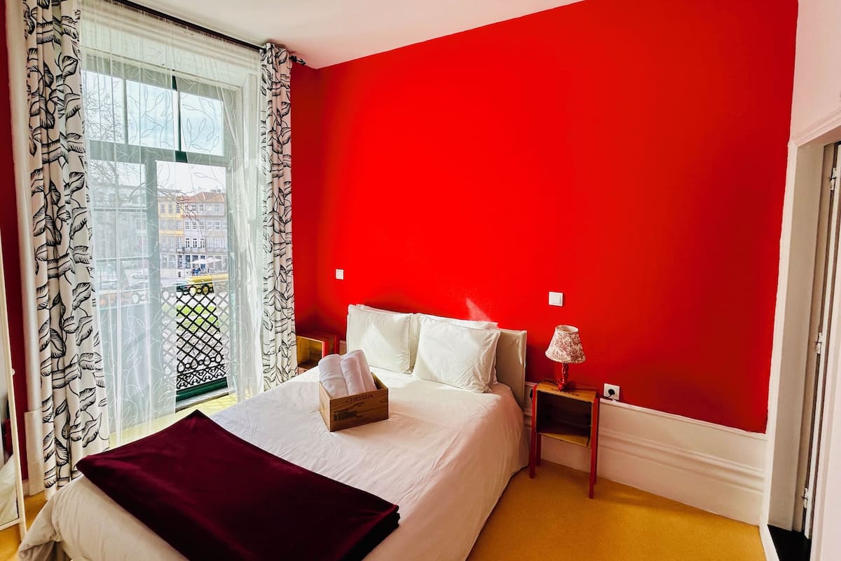 A bedroom with a red wall, a bed with white linens, a red throw blanket, a bedside table with a lamp, and a window with patterned curtains.