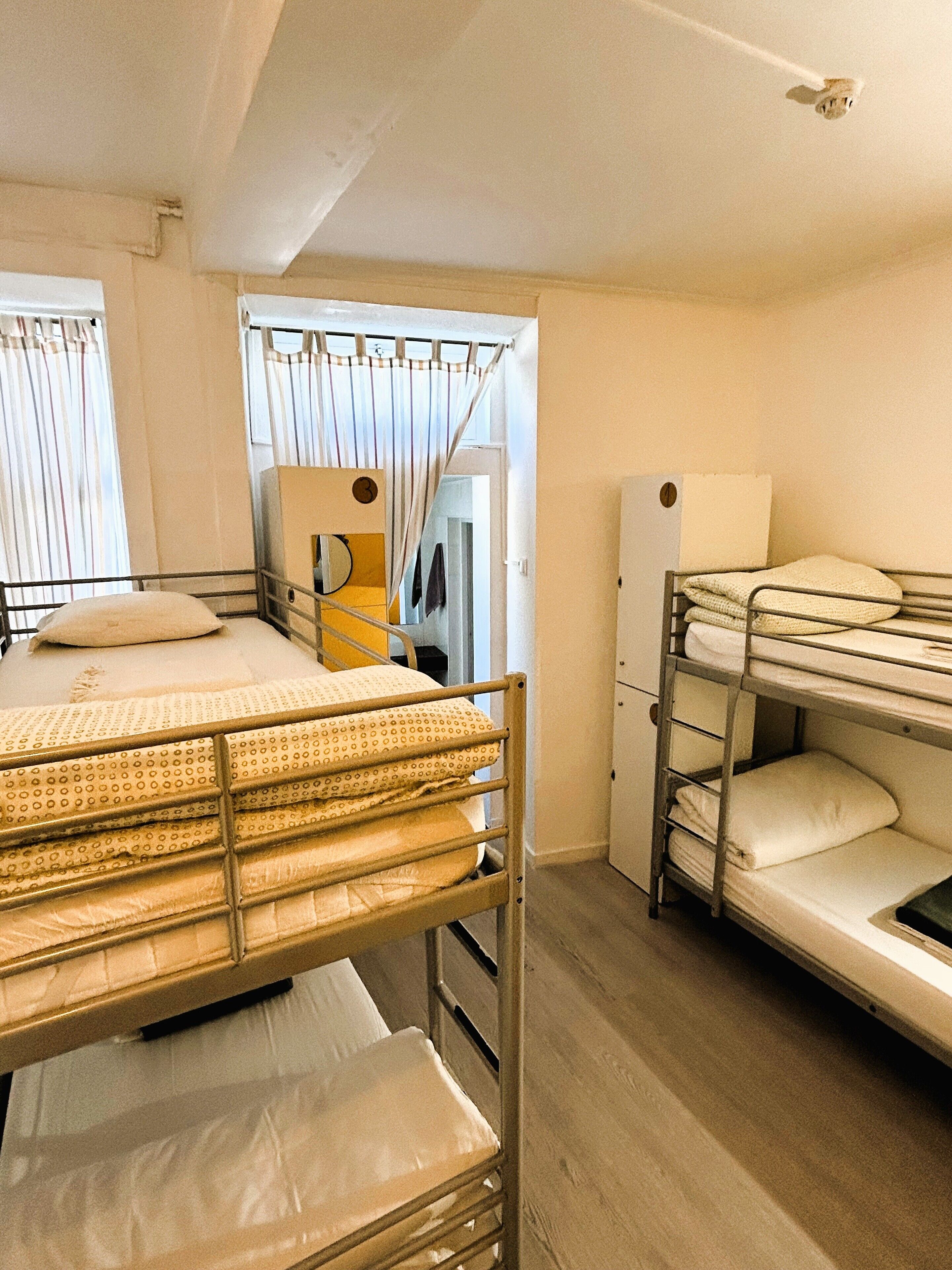 A dormitory room with bunk beds, a window with curtains, a mirror, and a wardrobe.