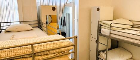 A dormitory room with bunk beds, a window with curtains, a mirror, and a wardrobe.