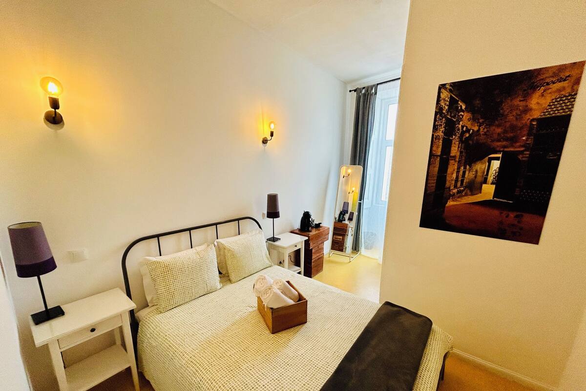 A neatly arranged bedroom with a bed, bedside tables, a lamp, a mirror, and a framed picture on the wall.