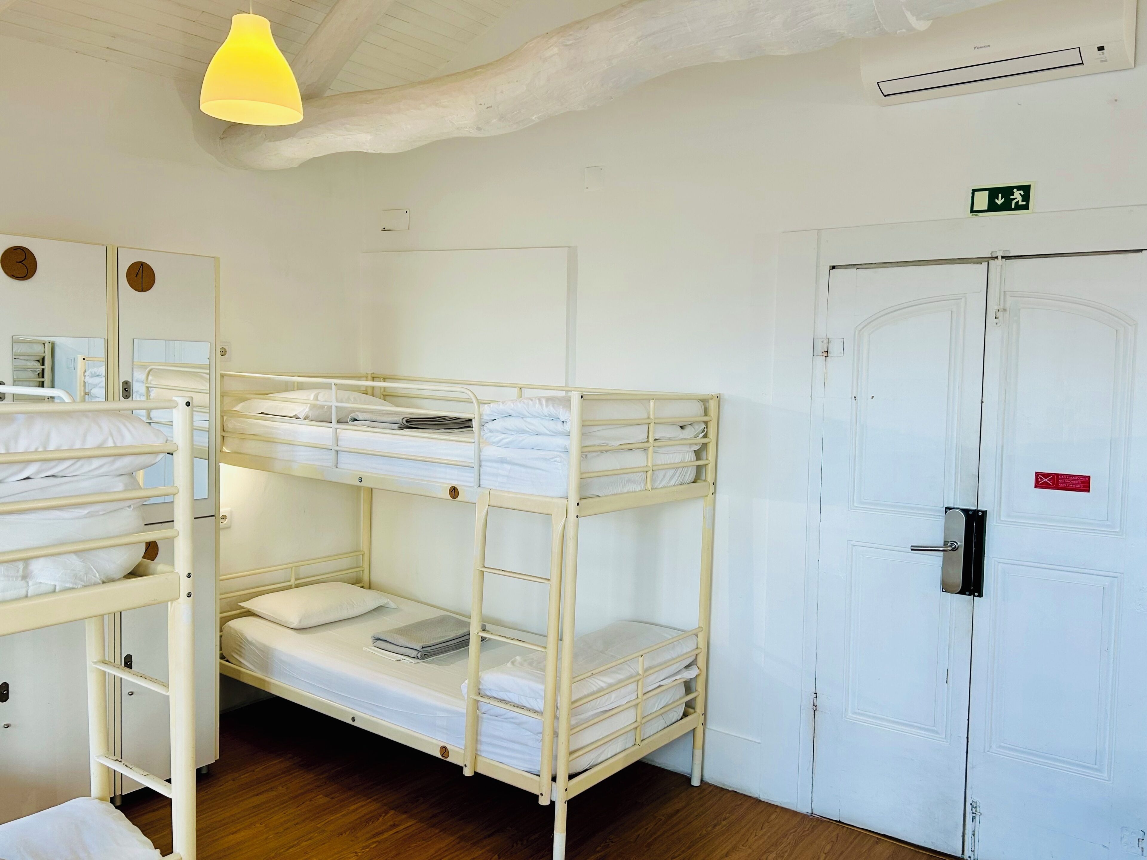 A dormitory room with bunk beds, a white door with an exit sign, and a ceiling light fixture.