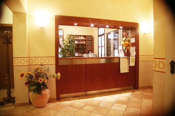 Una zona de recepción con un mostrador de madera, una planta grande en maceta con flores y una estantería al fondo.