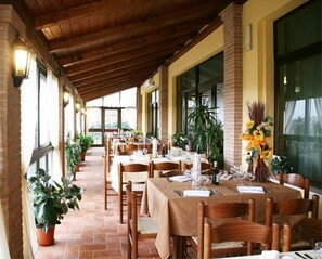 Un espacio de comedor al aire libre con techo, varias mesas y sillas, plantas en macetas y ventanas grandes.