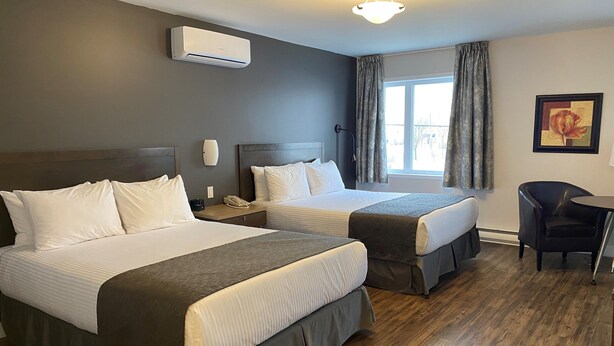 A hotel room with two beds, a window with curtains, a desk with a chair, and an air conditioning unit. - Hôtel Centre-Ville (Montmagny)