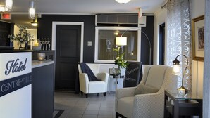 A hotel reception area with a reception desk, a white armchair, a sofa, a small table with a lamp, and a decorative vase with flowers. - Hôtel Centre-Ville (Montmagny)