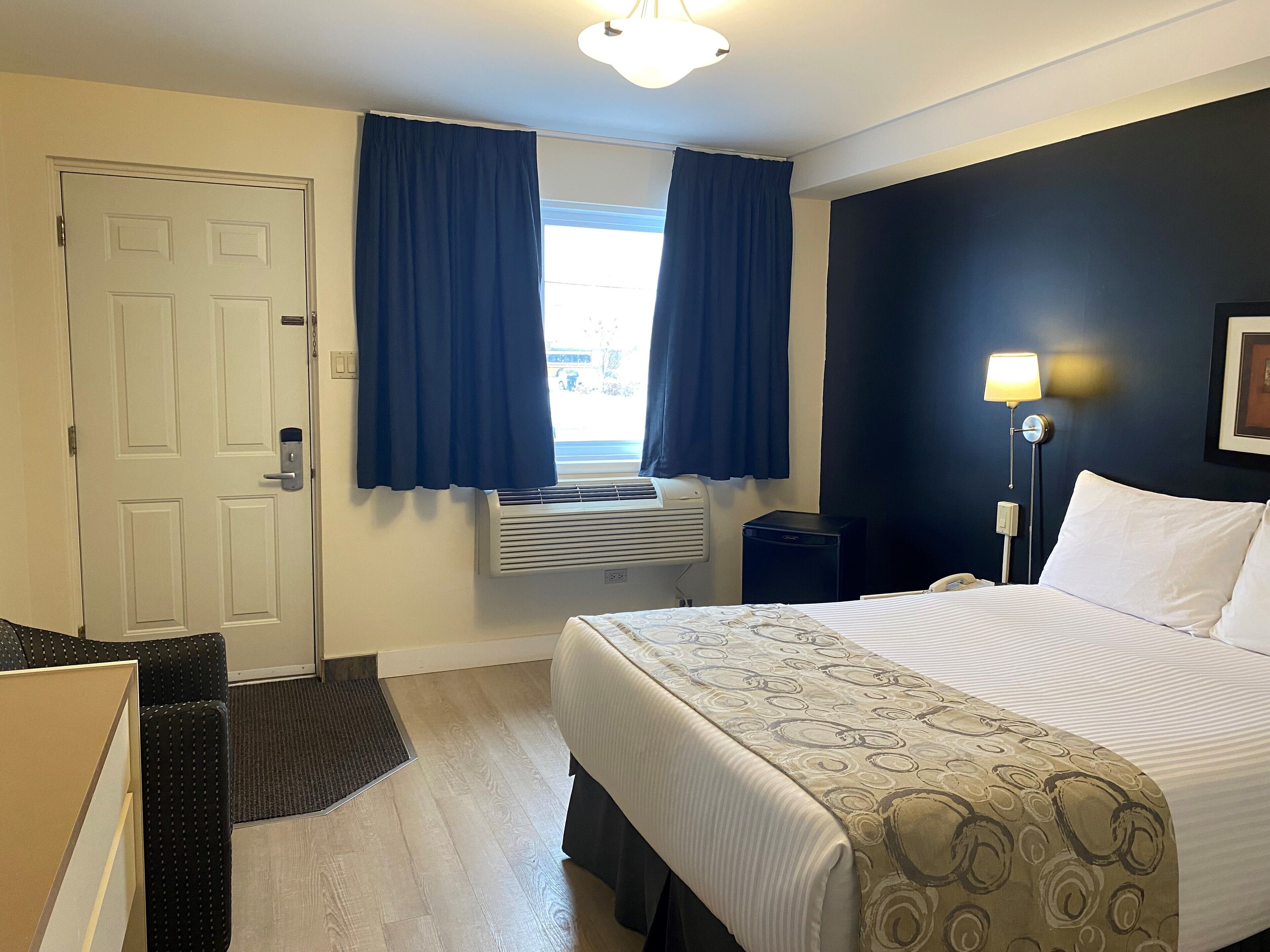 A hotel room with a bed, a desk, a chair, a desk lamp, a nightstand, a phone, a window with curtains, an air conditioning unit, and a door.