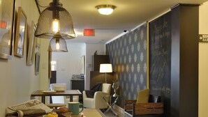 A cozy hotel room with a dining area featuring a table with a newspaper, a green mug, and a plate with food. There is a lamp, a chair, and a wall with a chalkboard menu. - Hôtel Centre-Ville (Montmagny)