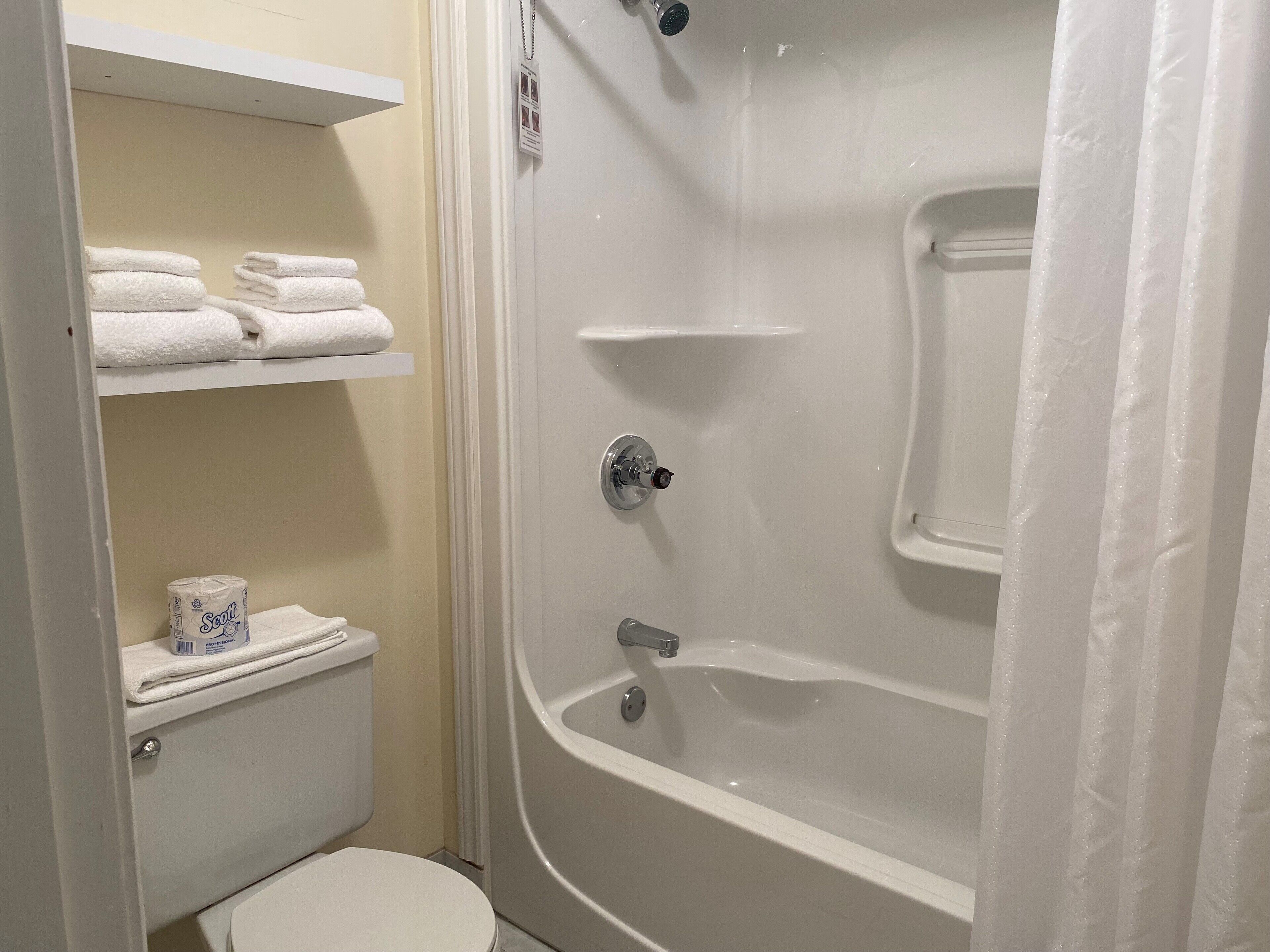 A bathroom with a toilet, a shower, and a sink. There are towels and toilet paper on the shelves.