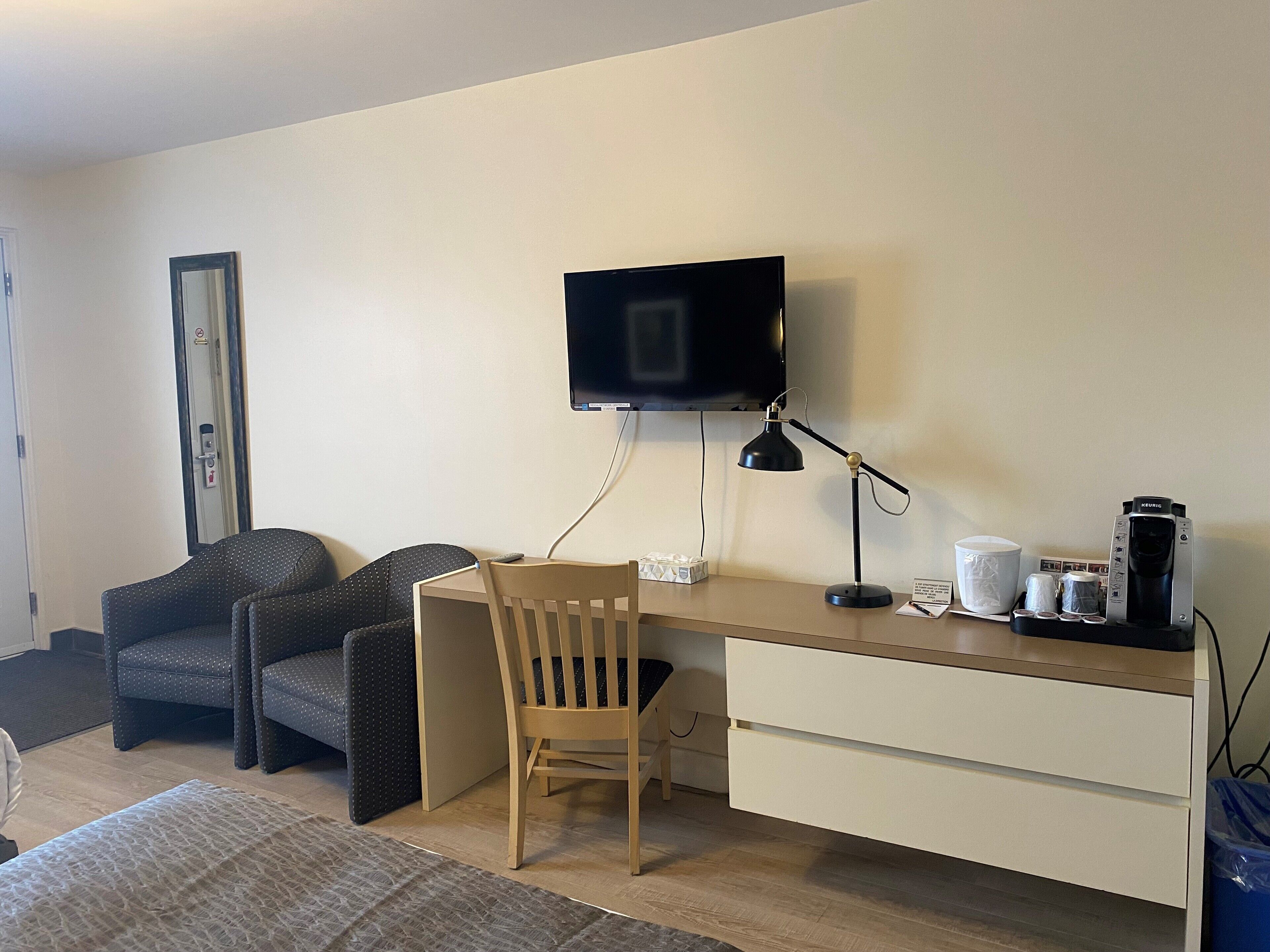 A hotel room with a wooden desk, a television mounted on the wall, a black desk lamp, a coffee maker, and two upholstered chairs.