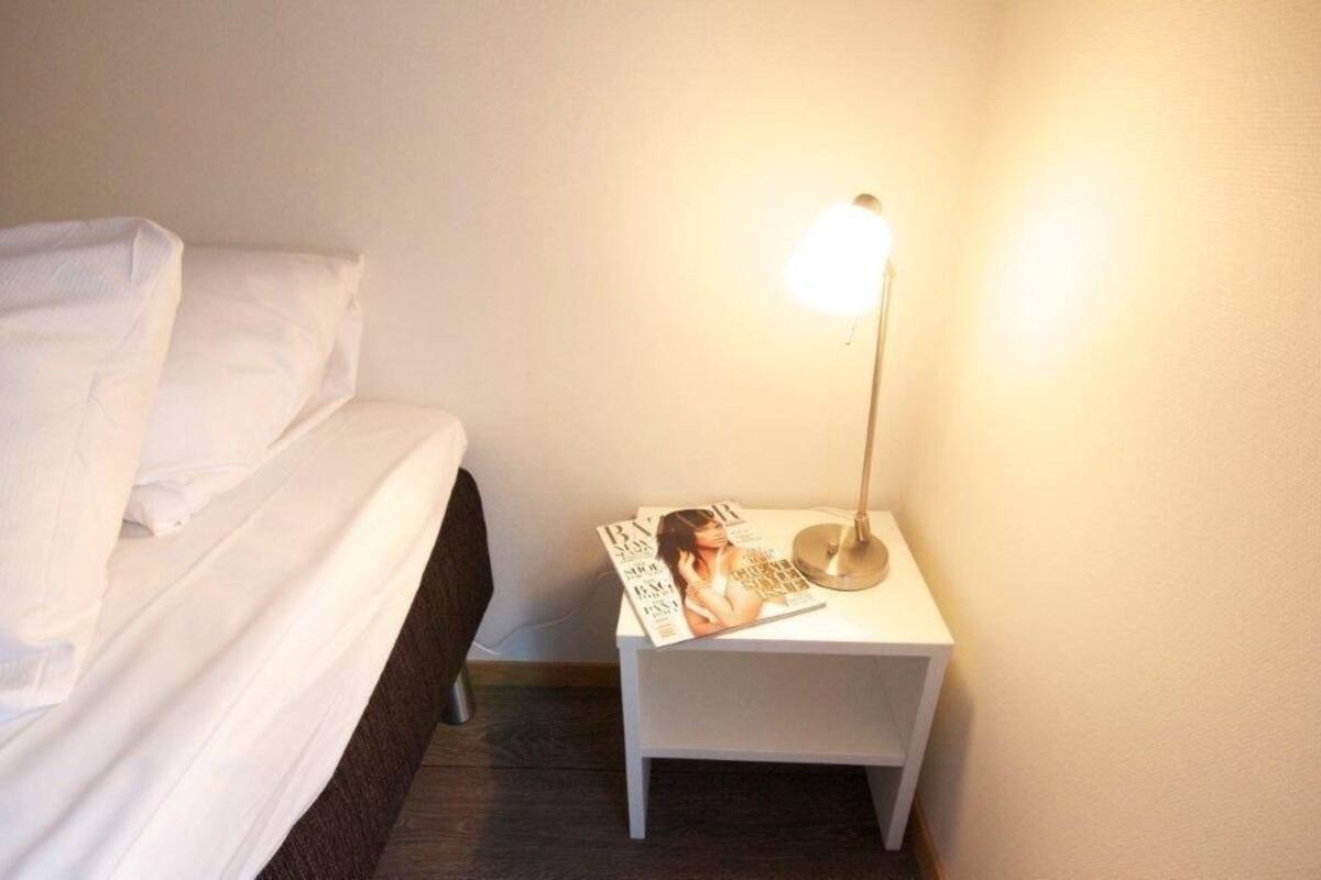 Ein ordentlich bezogenes Bett mit weißen Leinen, ein Nachttisch mit einer Zeitschrift und eine Stehlampe, die Licht spendet.