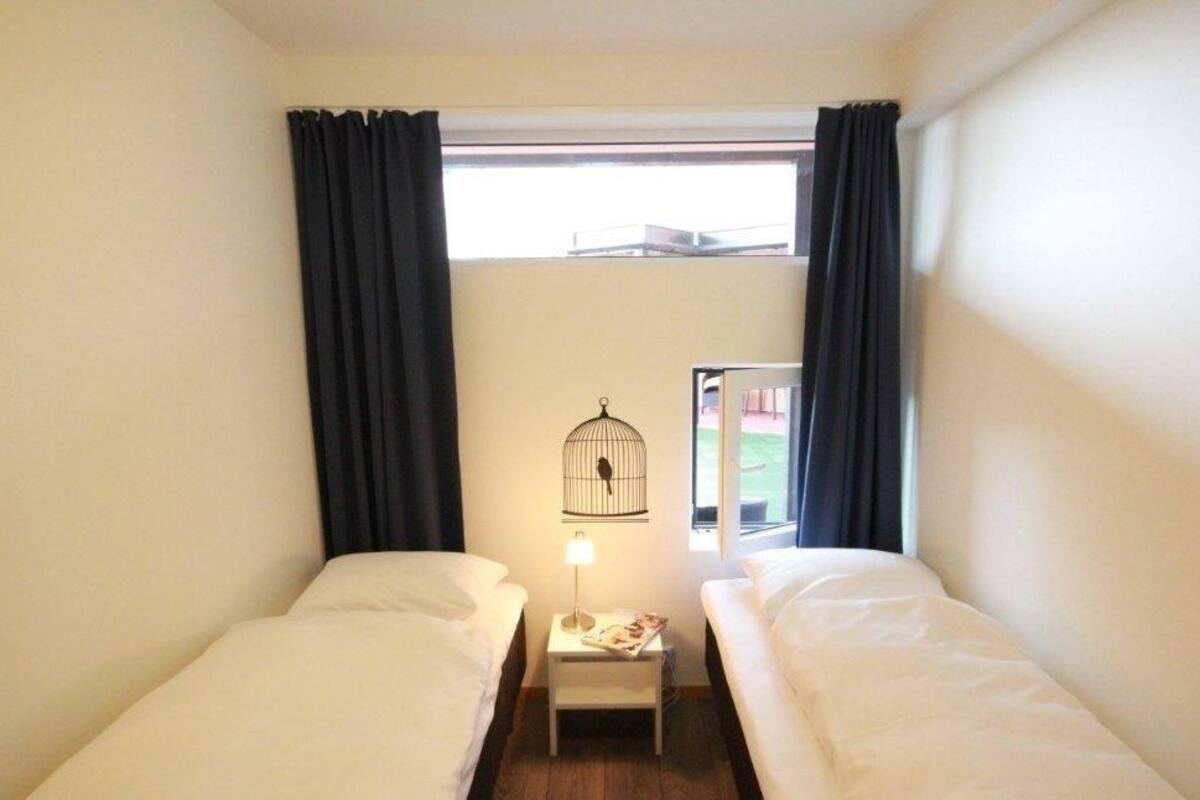 Ein kleines Hotelzimmer mit einem Bett, einem Nachttisch mit Lampe, einem Fenster mit Vorhängen und einem Vogelkäfig an der Wand.