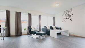 Una sala de estar moderna con sofá, sillón y una mesita de centro. Cuenta con ventanas grandes con cortinas, una planta y un aplique de pared.