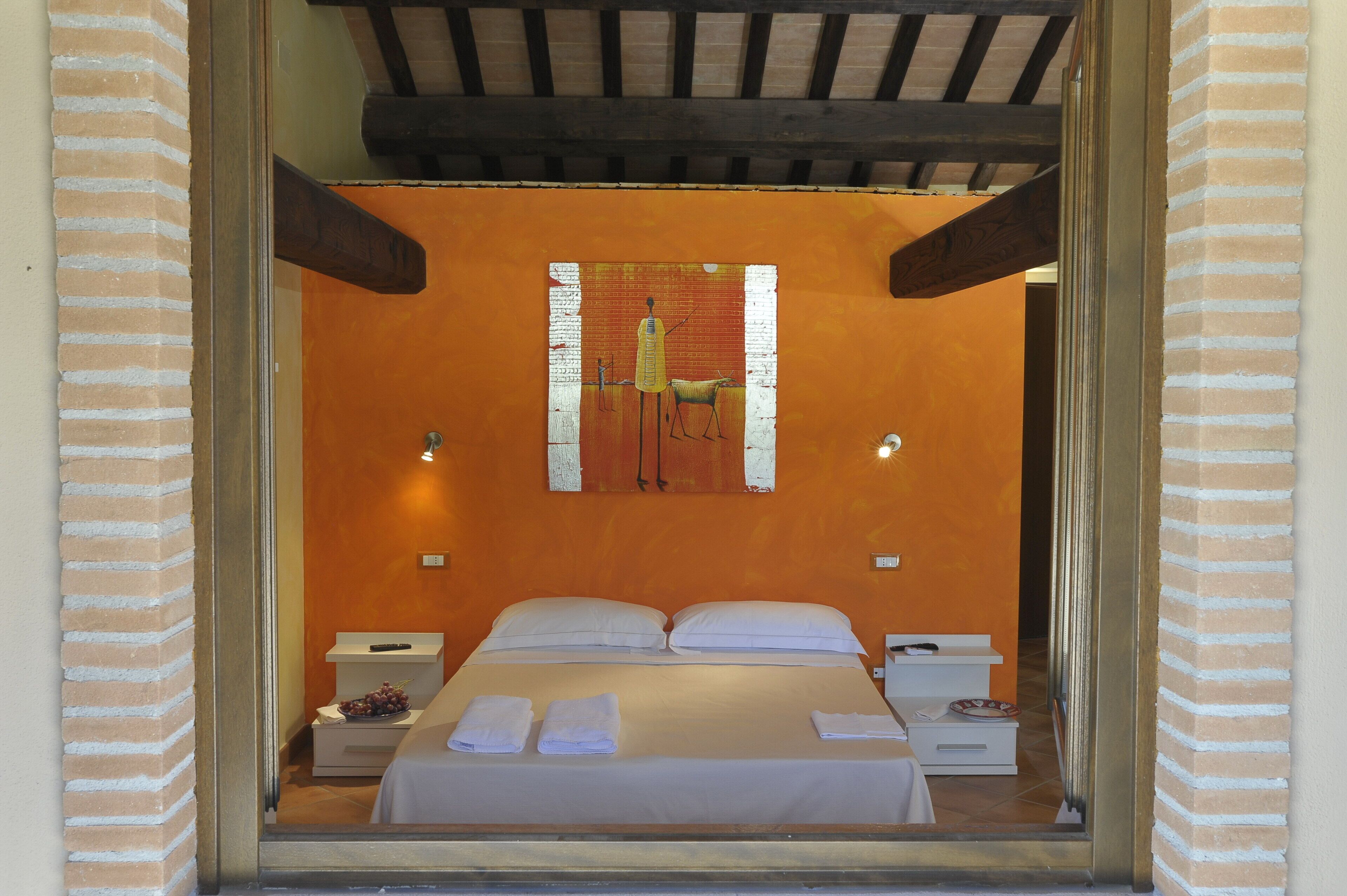 Una habitación de hotel con una cama, almohadas blancas y una mesita de noche con una lámpara y un plato de uvas.