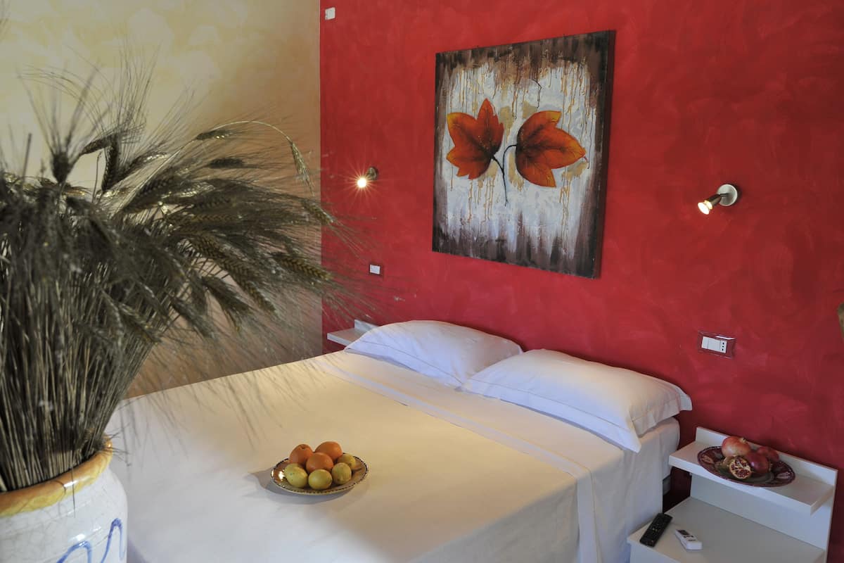 A hotel room with a bed, a bowl of fruit, a potted plant, and a painting on the wall.