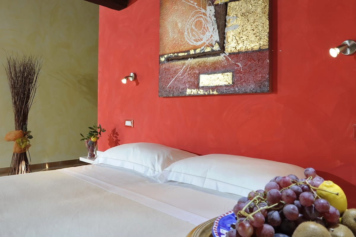 A bedroom with a red wall, a bed with white linens, a vase with fruits, and a painting.