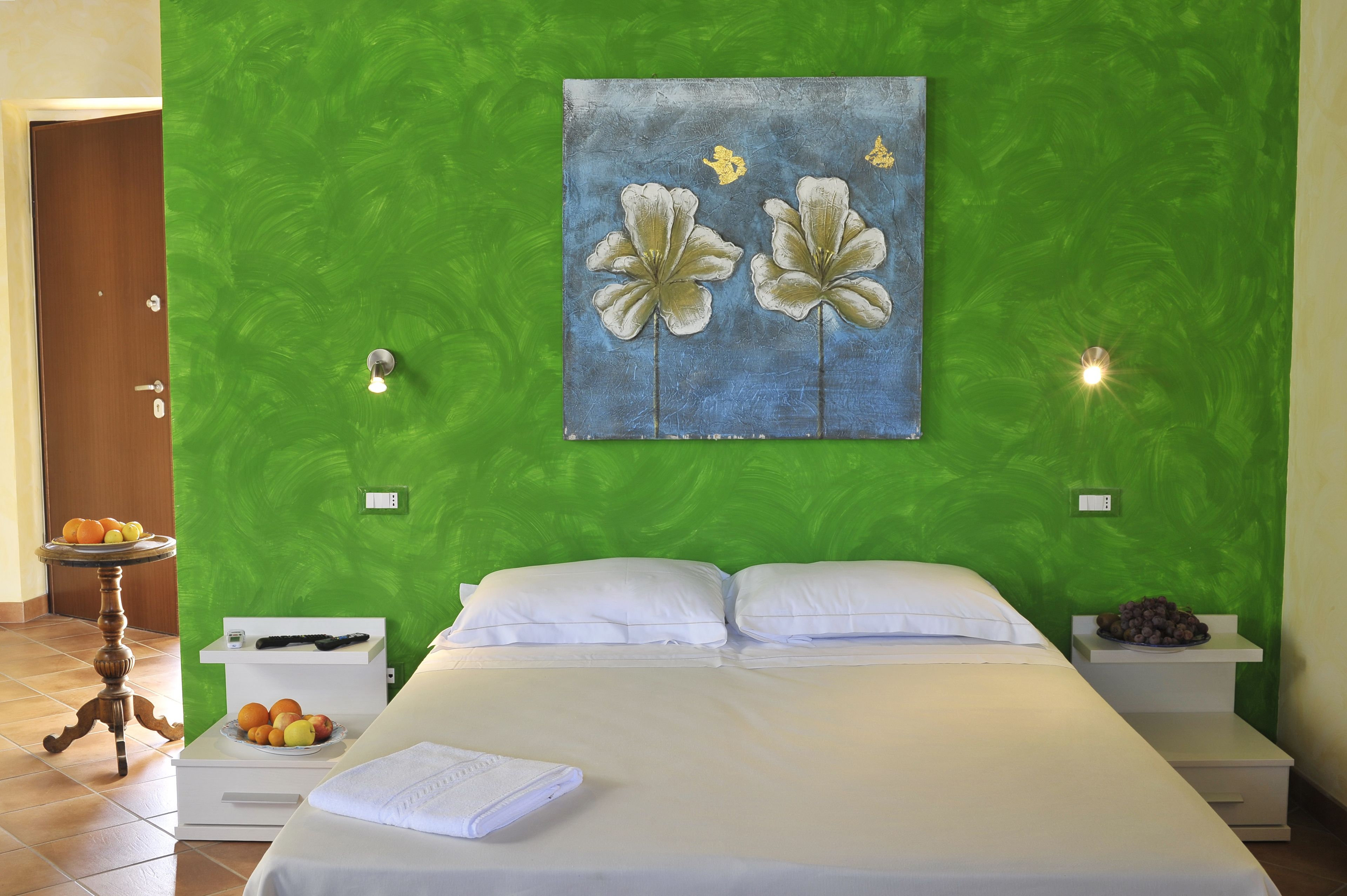 A hotel room with a green wall, a bed with white linens, two bedside tables with fruit bowls, and a painting of flowers.