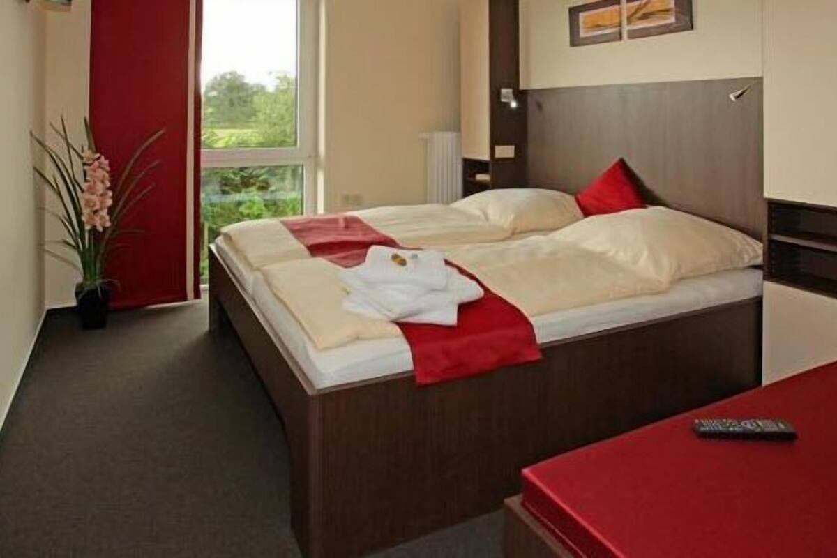 Un lit double avec une couverture rouge et des oreillers, une table de chevet avec une télécommande, et une vue sur la verdure à l’extérieur.