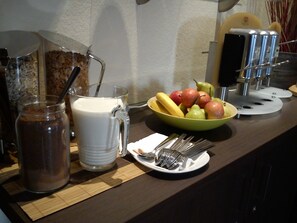 Preparación para el desayuno con un tazón de fruta, una taza, un tarro de café y un plato de cubiertos.