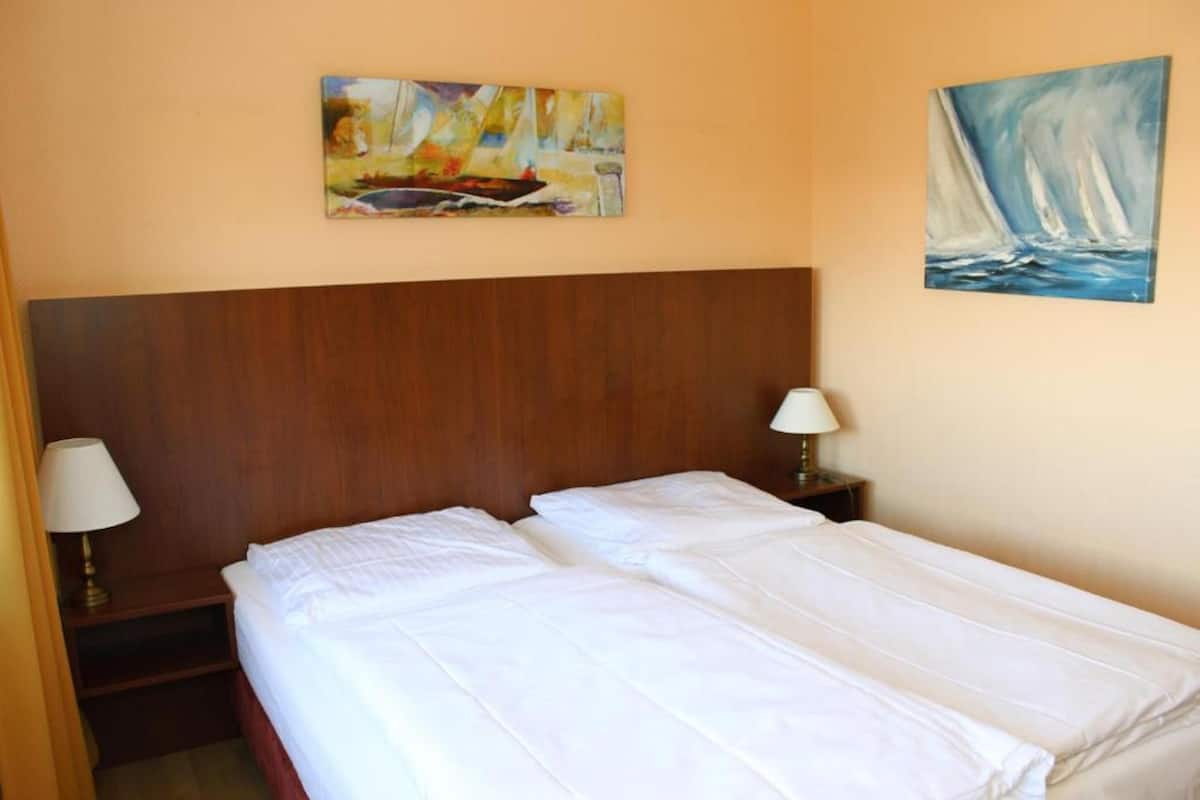A double bed with white linens, a wooden headboard, two bedside tables with lamps, and two paintings on the wall.