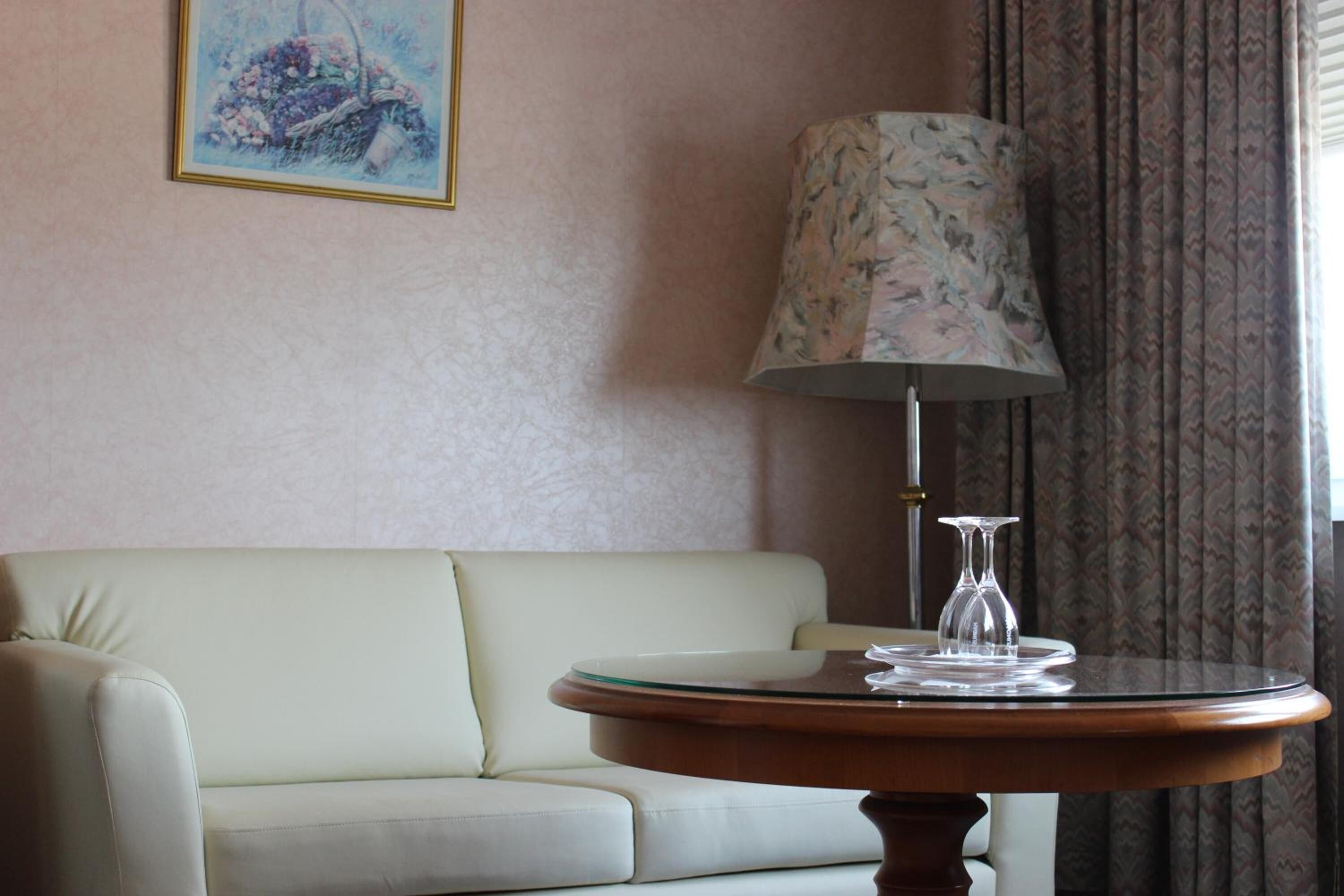 A room with a light-colored sofa, a round wooden table with a glass top, a floral lampshade, and a framed artwork on the wall.