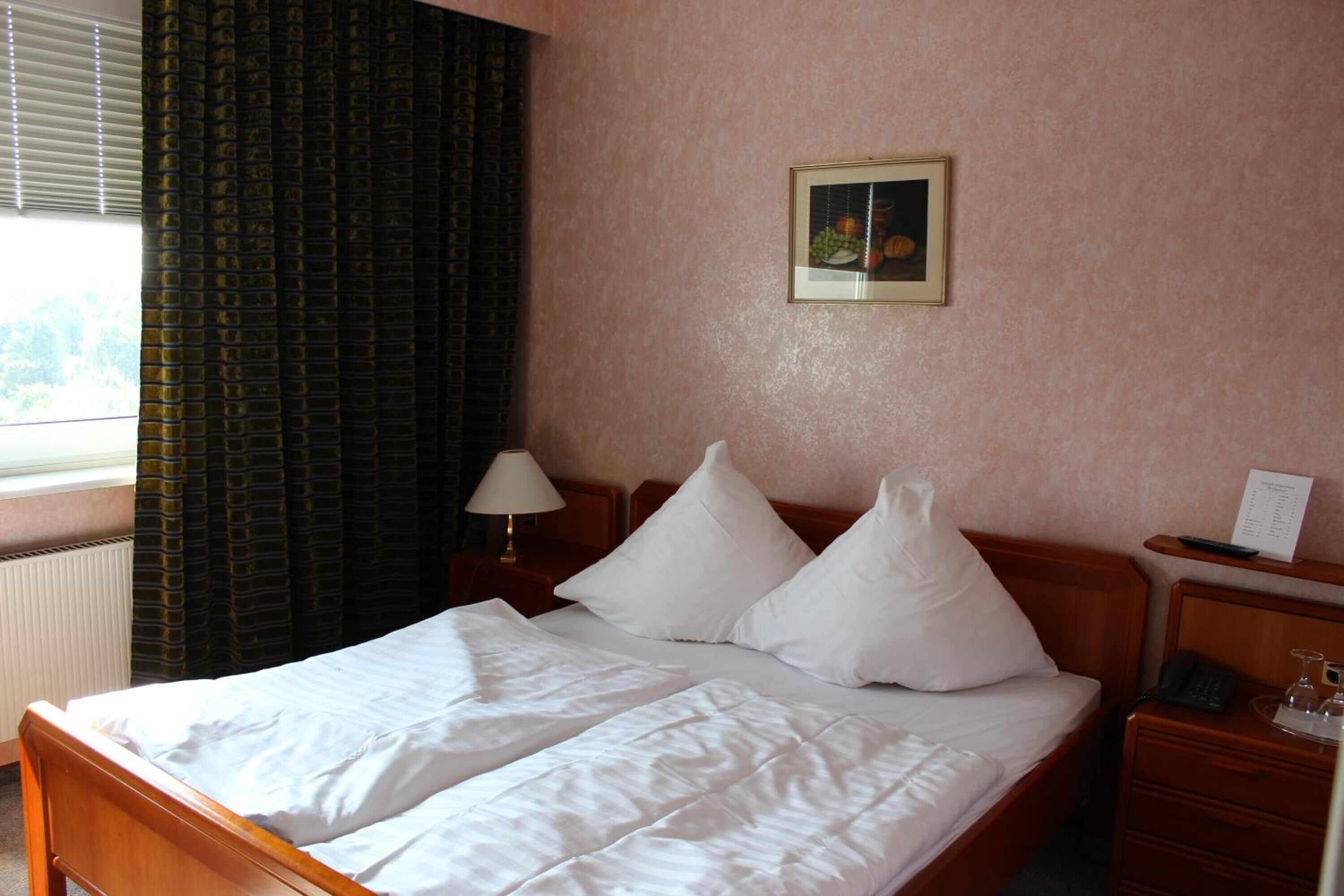 Ein Hotelzimmer mit Bett, Nachttisch, Lampe, einem gerahmten Bild an der Wand und einem Fenster mit Vorhängen.