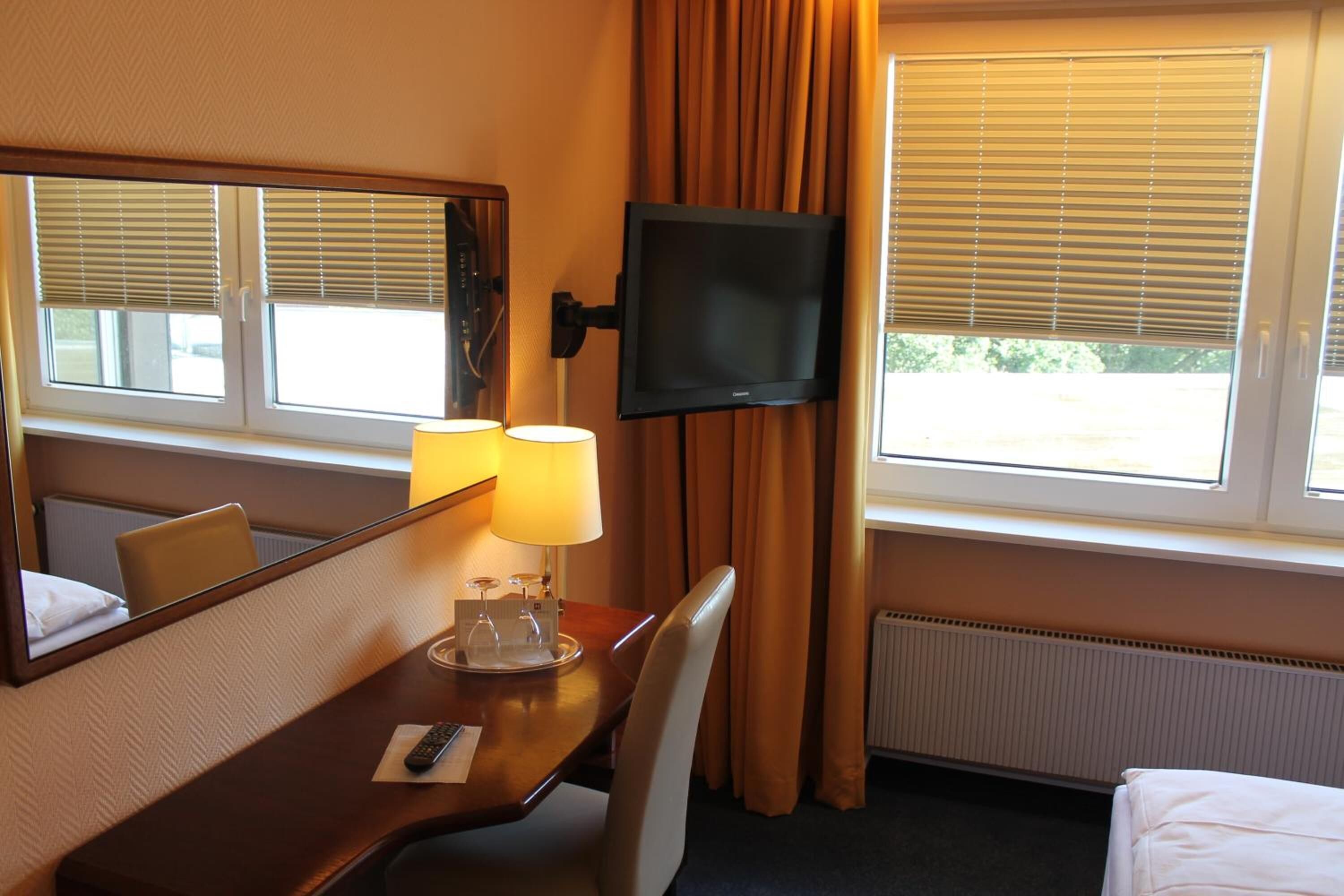 Et hotelværelse med seng, skrivebord med lampe, fjernsyn og vinduer med gardiner.