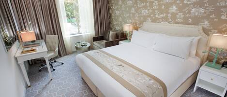A hotel room with a large bed, a desk with a chair, a small table, and a nightstand with a lamp.