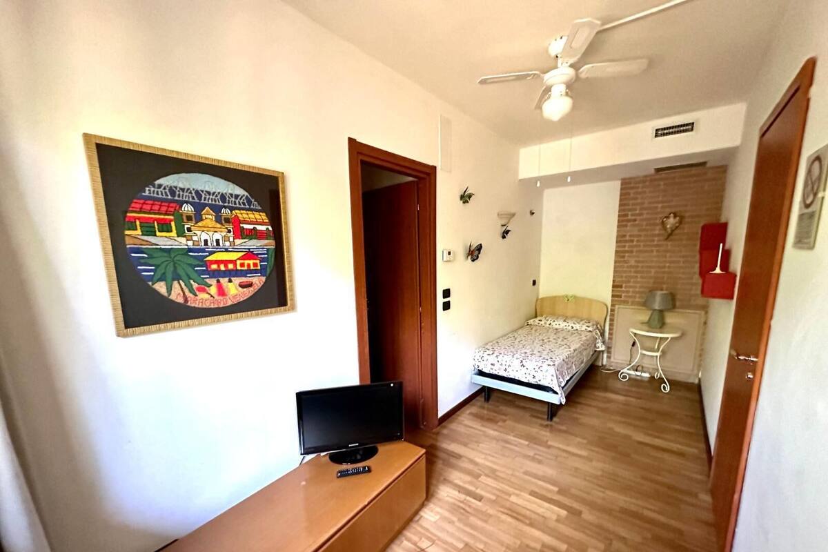A room with a bed, a desk with a TV, a ceiling fan, and a wall with a colorful painting.