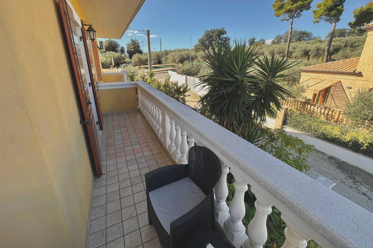 A balcony with a tiled floor, a wicker chair, and a view of a residential area with trees and a house.