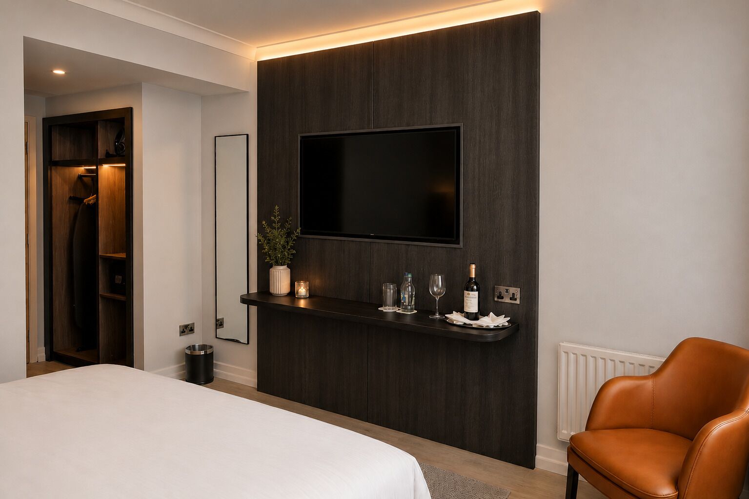 A modern hotel room with a bed, a seating area with a leather chair, a mounted TV, and a small bar area with glasses and a bottle.