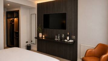 A modern hotel room with a bed, a seating area with a leather chair, a mounted TV, and a small bar area with glasses and a bottle.