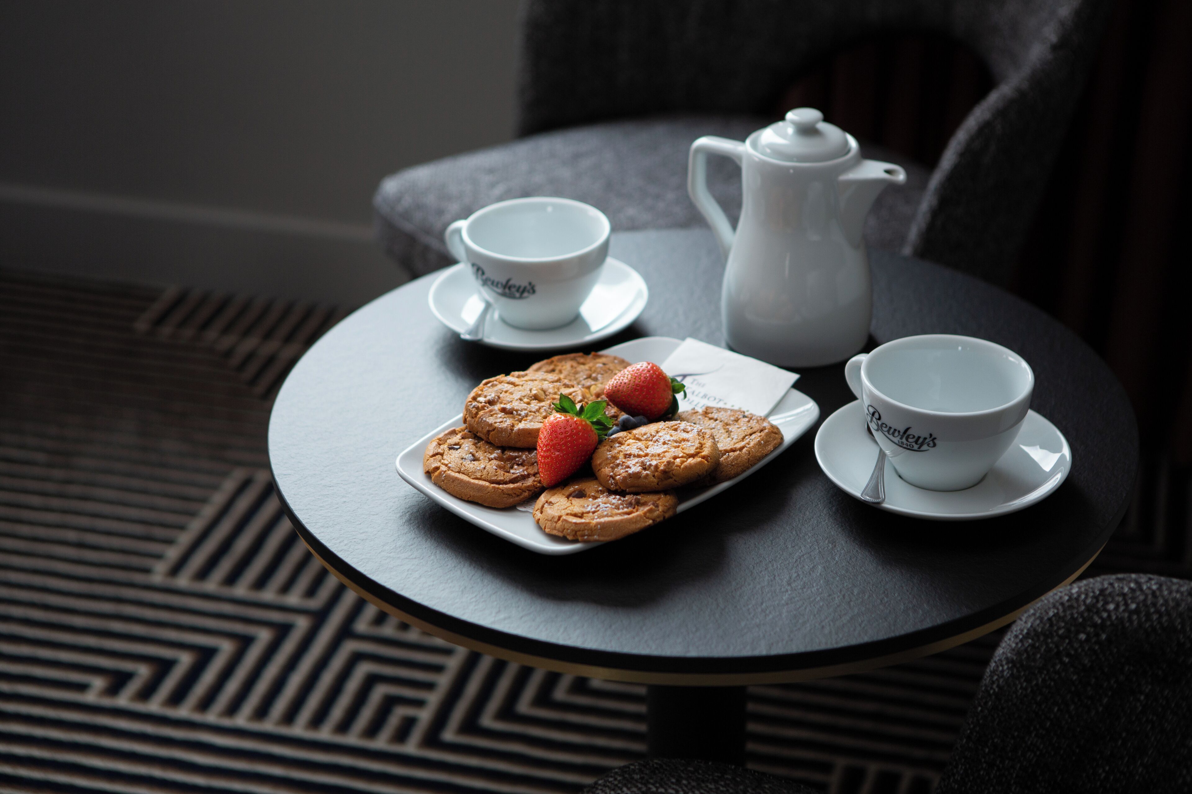 Une table ronde sur laquelle se trouvent un plateau de biscuits et des fraises, deux tasses à thé et une théière, le tout posé sur un sol à motifs.