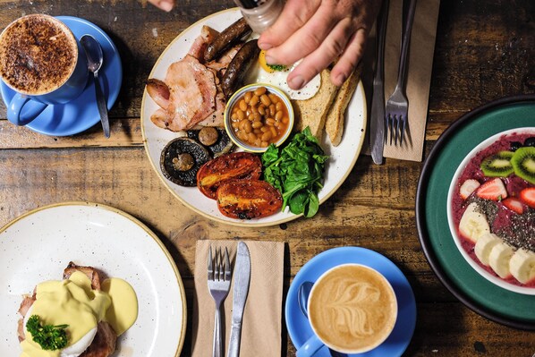 Una mesa de desayuno con diversos platos, incluyendo huevos, frijoles, tocino y tostadas, junto con tazas de café y té.