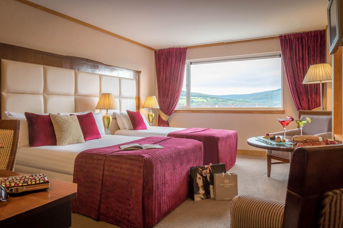 A hotel room with a large bed, two bedside tables, a small round table with drinks, a sofa, and a view of the countryside.