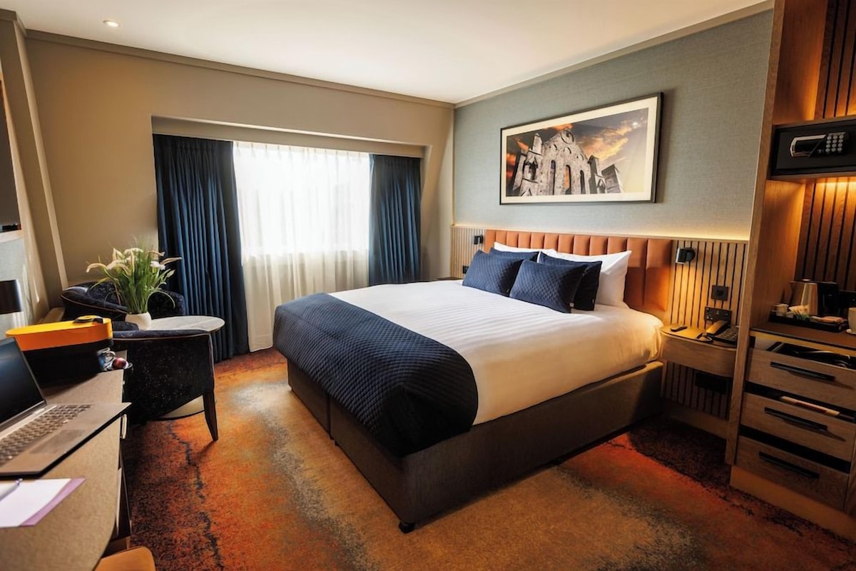 A hotel room with a large bed, a desk with a laptop, a bedside table with a lamp, and a painting on the wall.