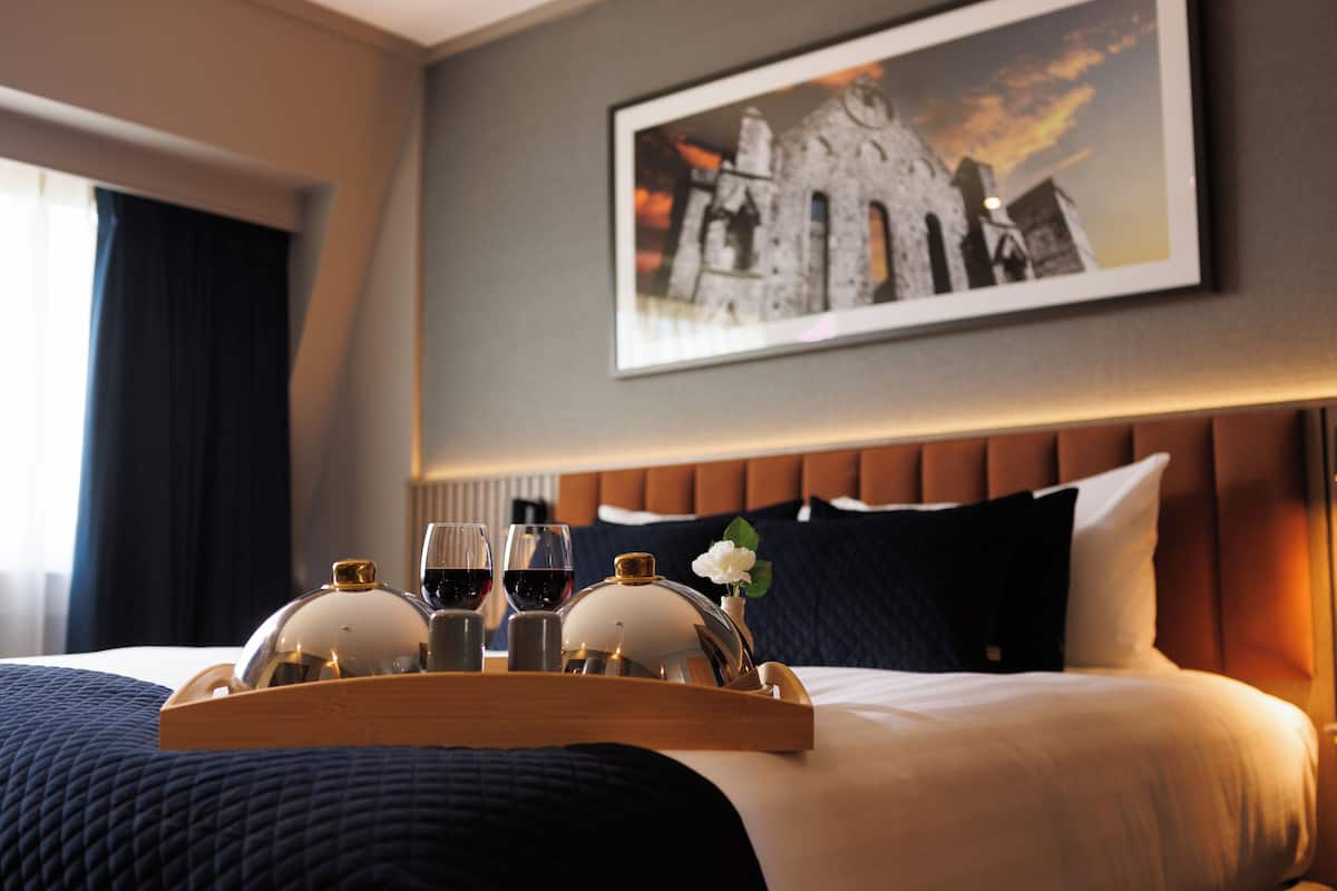 A neatly made bed with a tray of glasses and a bottle, a framed picture on the wall, and a window with curtains.
