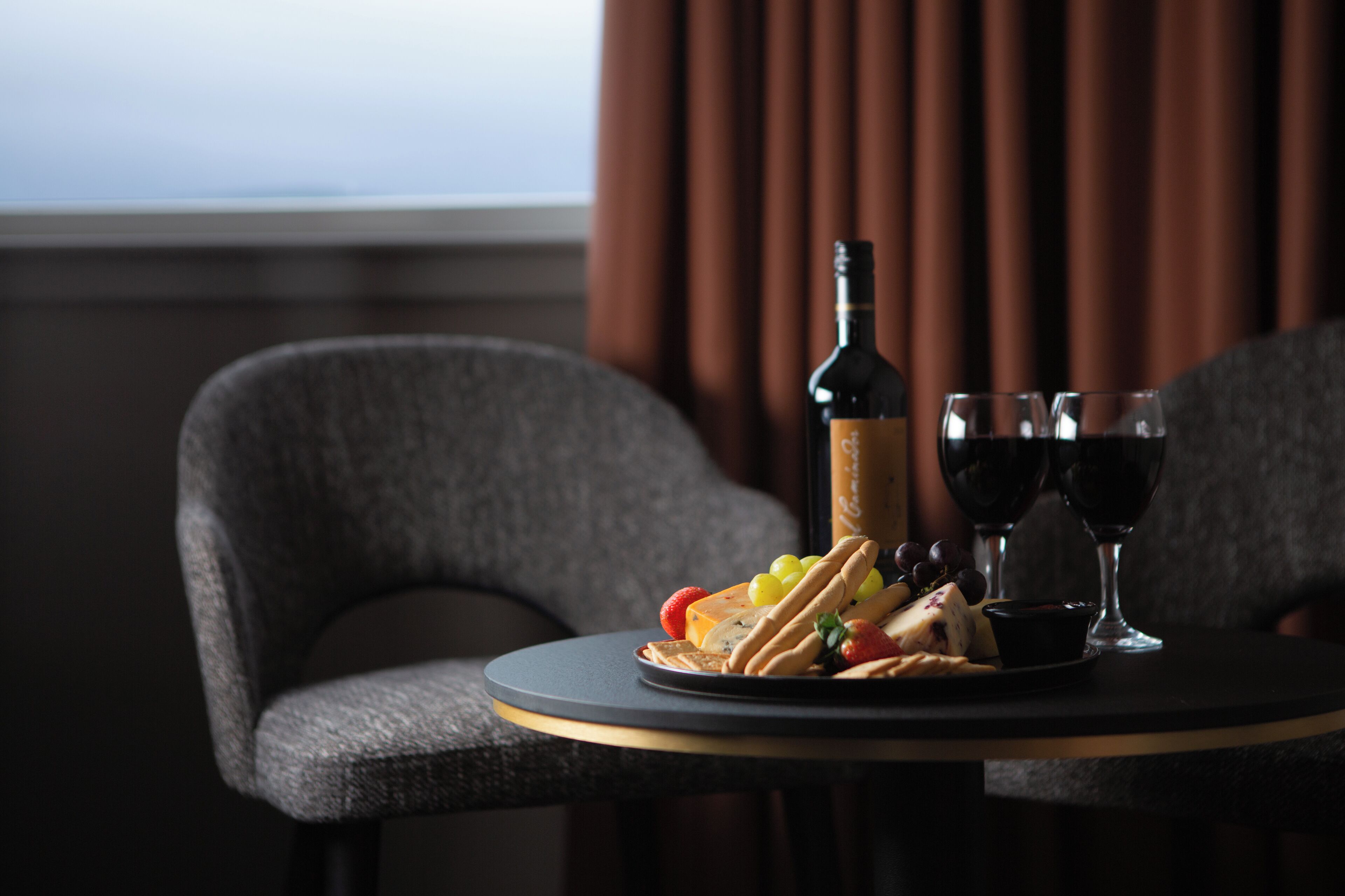 Une table avec une bouteille de vin, deux verres et une assiette de fromage, de crackers et de raisins.