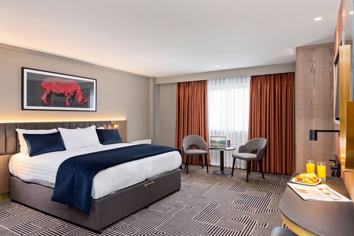 A hotel room with a large bed, a desk with a chair, a small table with a chair, a framed picture on the wall, and a window with curtains.