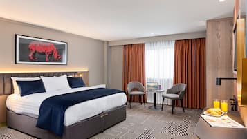 Habitación de hotel con una cama grande, un escritorio con silla, una mesita con otra silla, un cuadro en la pared y una ventana con cortinas.