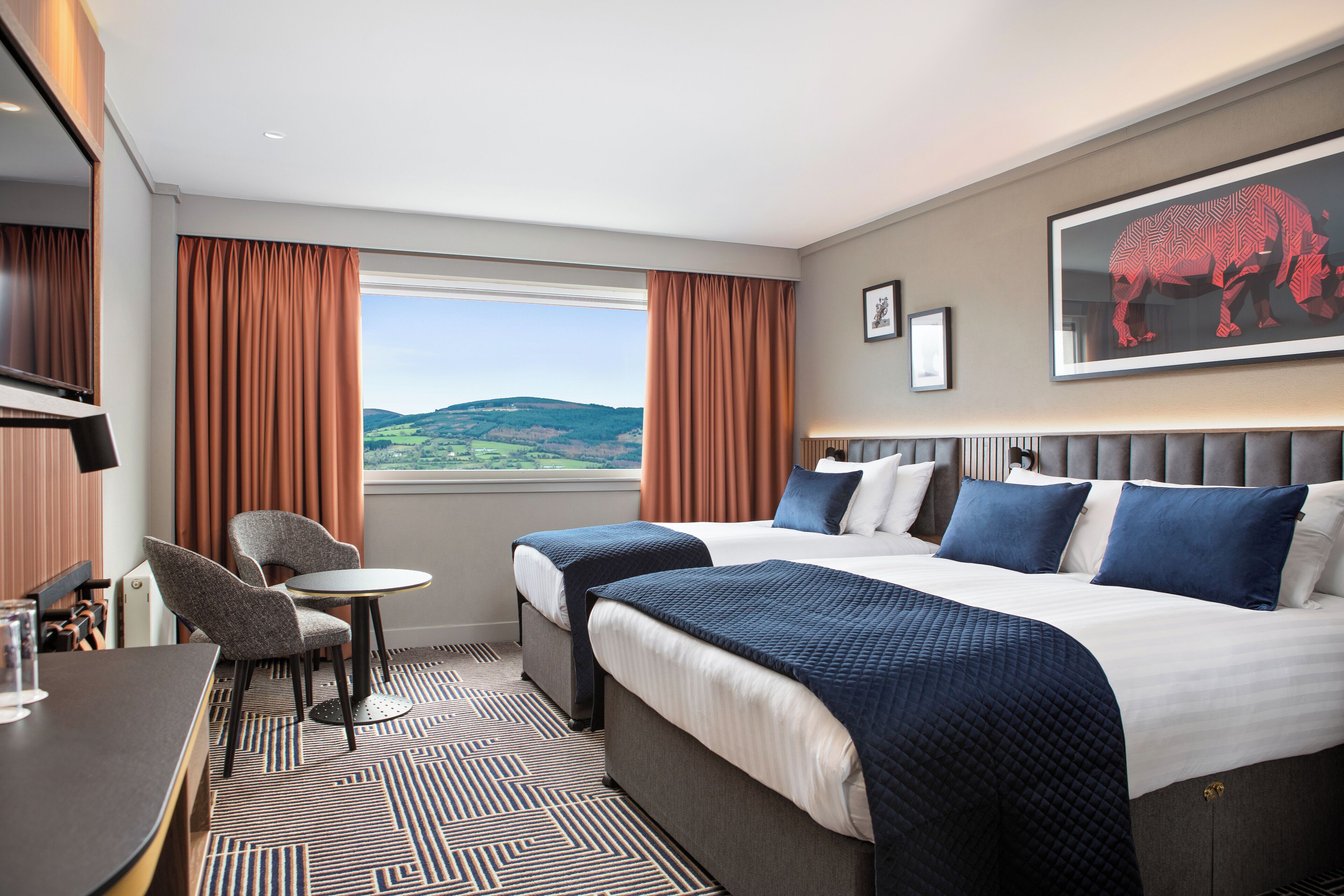 Habitación de hotel con dos camas, un escritorio, una silla y un ventanal con vistas a las colinas.