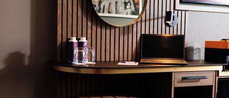 A hotel room with a wooden panel wall, a round mirror, a laptop, a coffee maker, and a cushioned chair.
