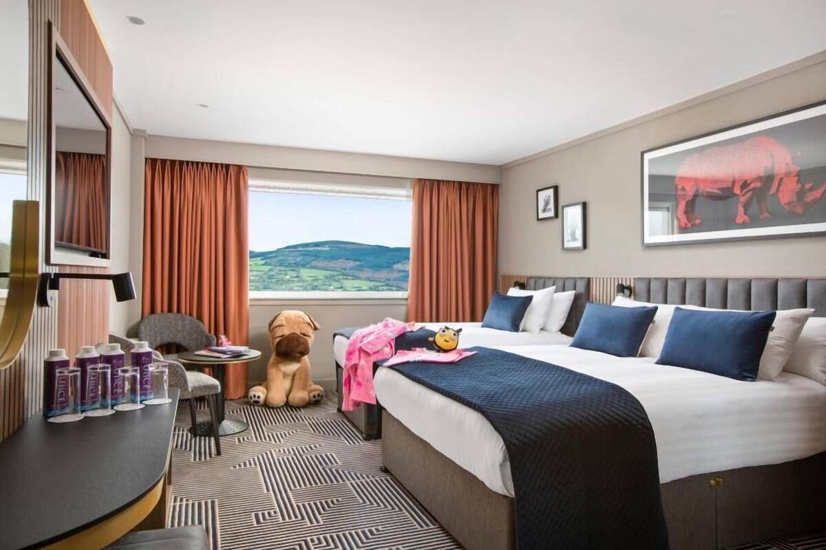 A hotel room with a large bed, a desk with a chair, a window with curtains, a dog, and a view of the countryside.