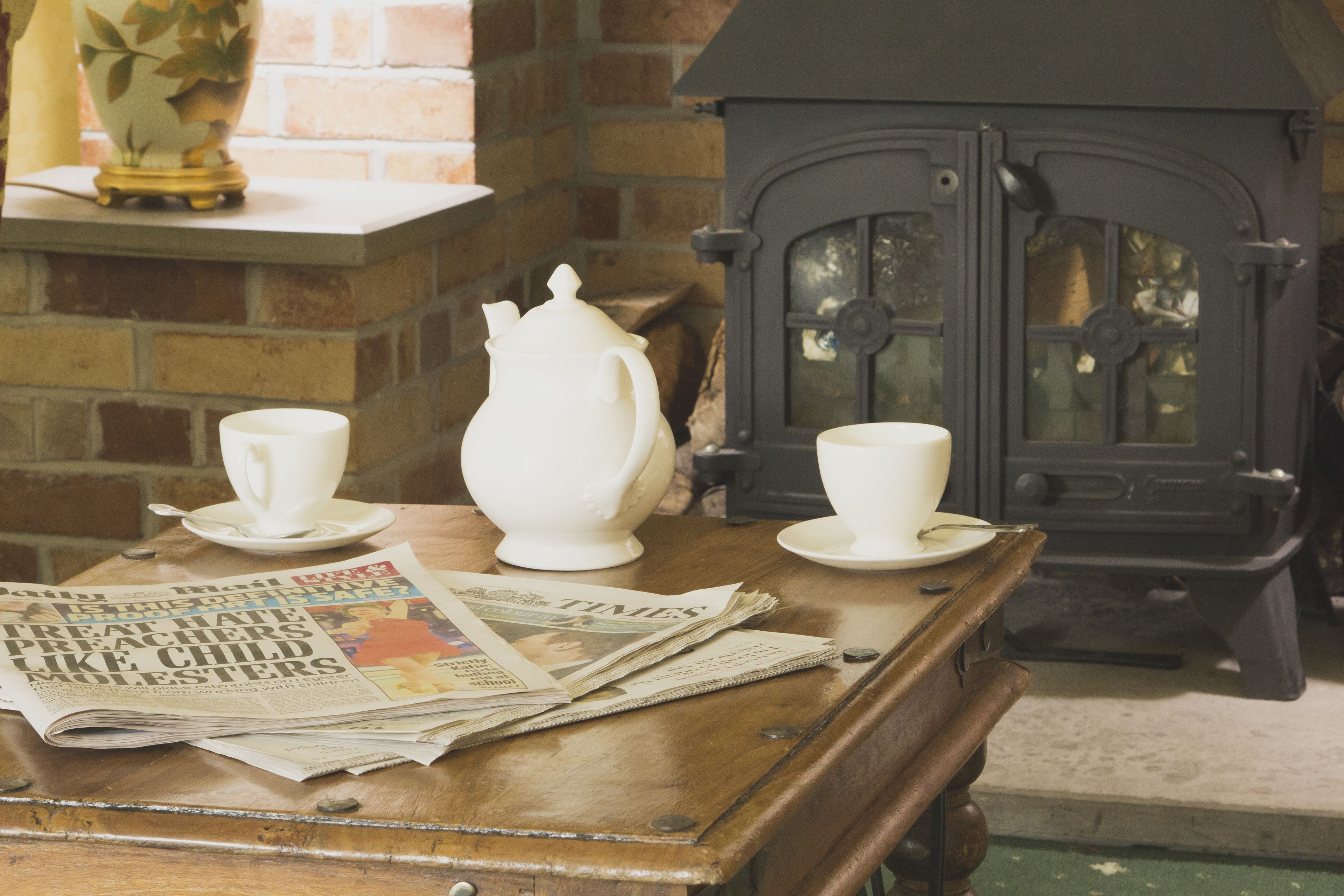A wooden table with a newspaper, a white teapot, and two cups.