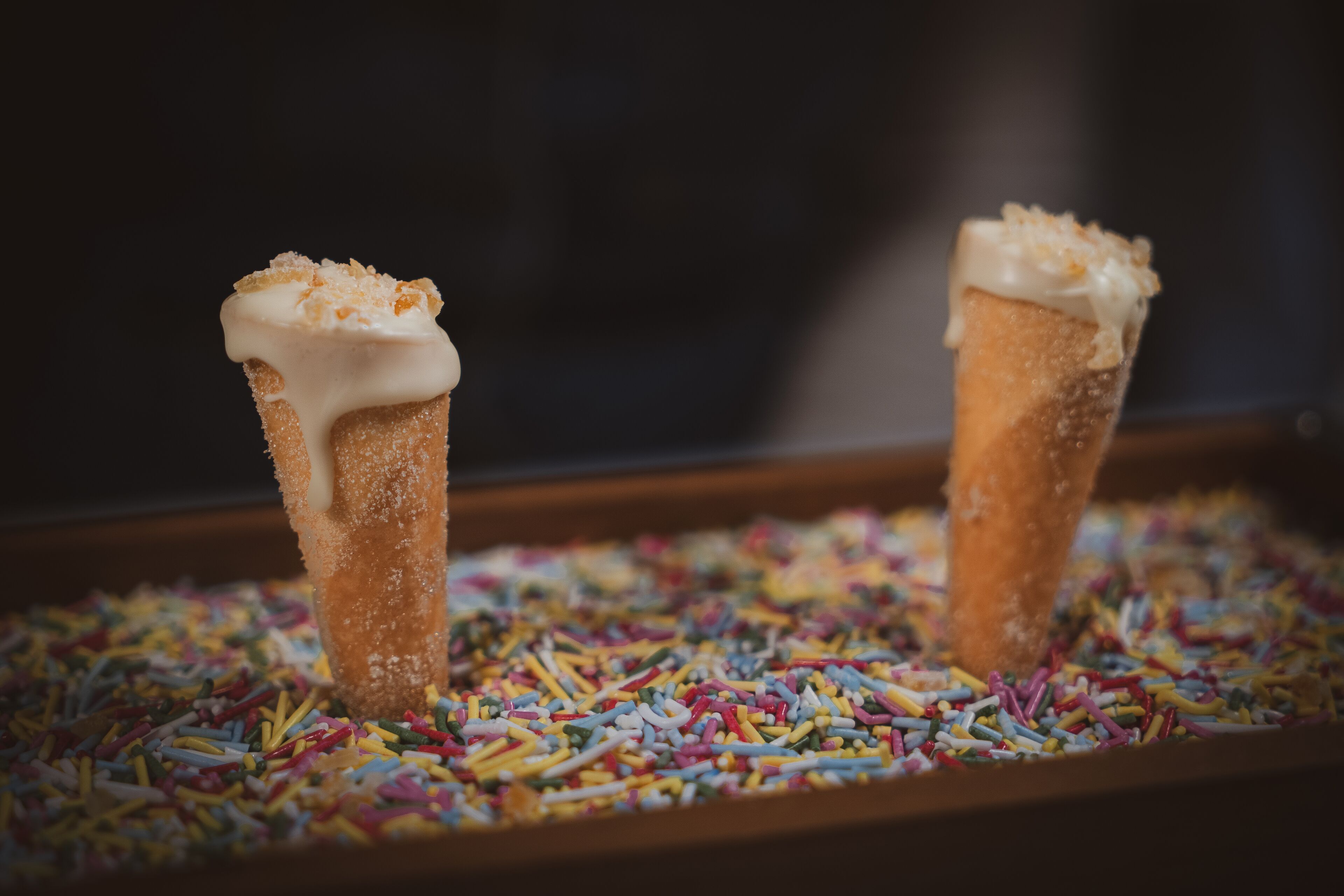 Two sugar cones with white icing and toppings on a tray of colorful sprinkles.