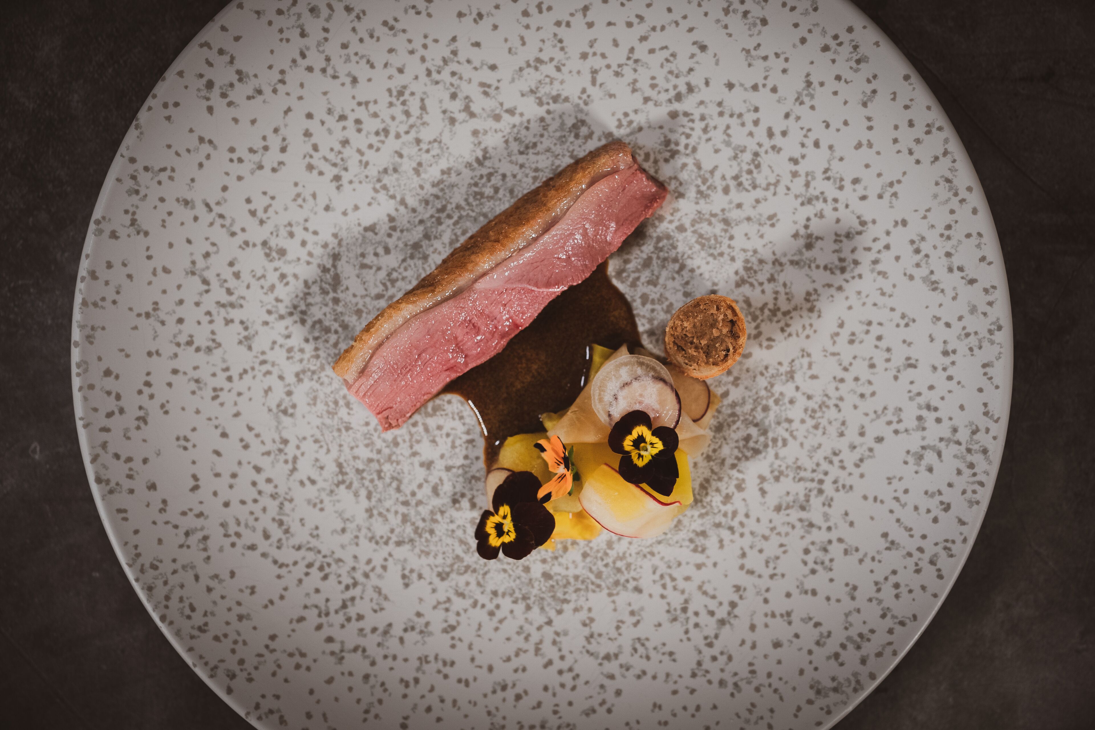 A round plate with a piece of meat, a slice of bread, and some fruit and flowers.